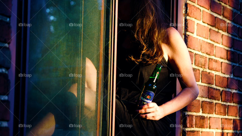 City life Woman sitting in window of brick apartment or house to cool off on hot summer day drinking refreshing Heineken 0.0 with golden hour sun and reflection urban lifestyle photography part of a series 