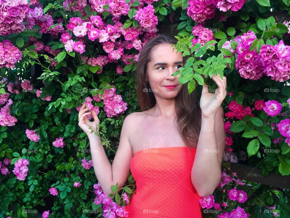 Portrait of pretty woman in red dress standing against wall of pink roses