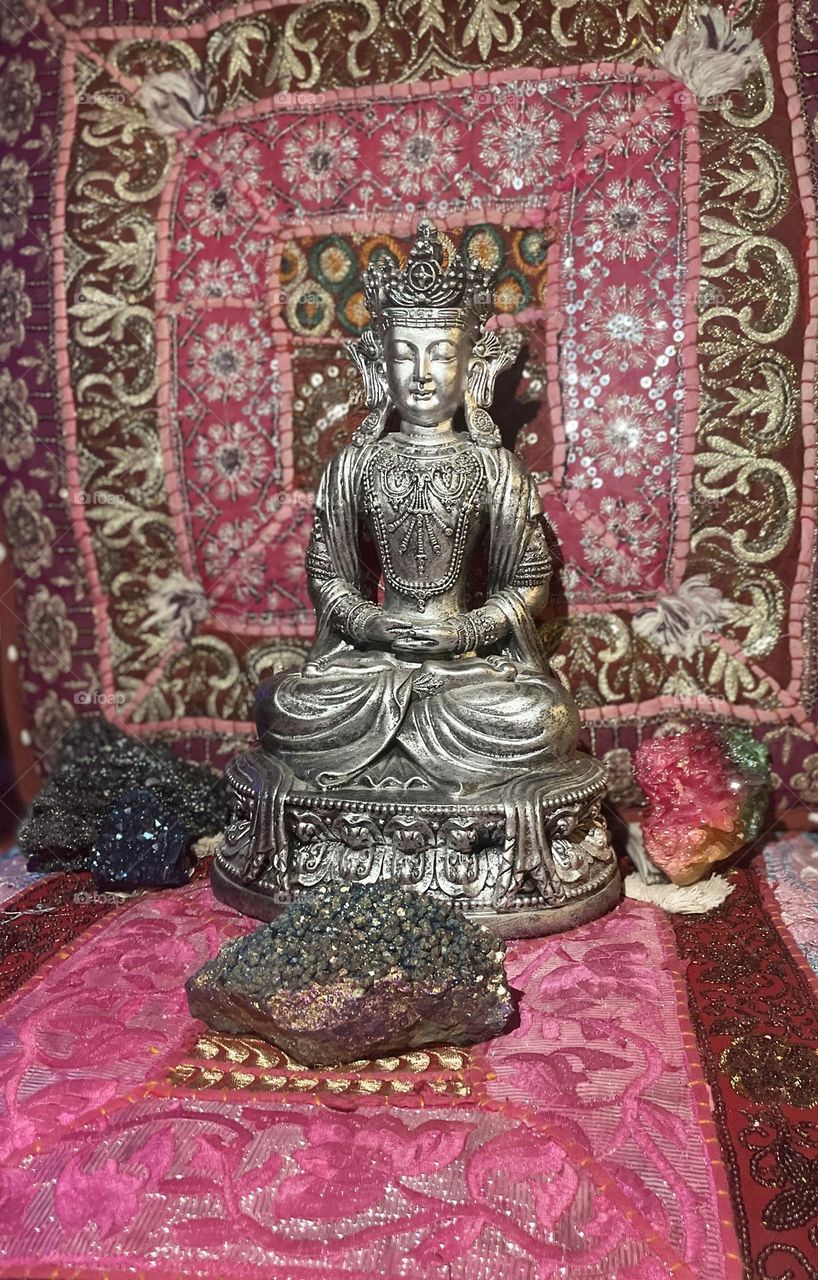 A silver and black Buddha meditating amongst several beautiful and colorful crystals with a nice colorful background.