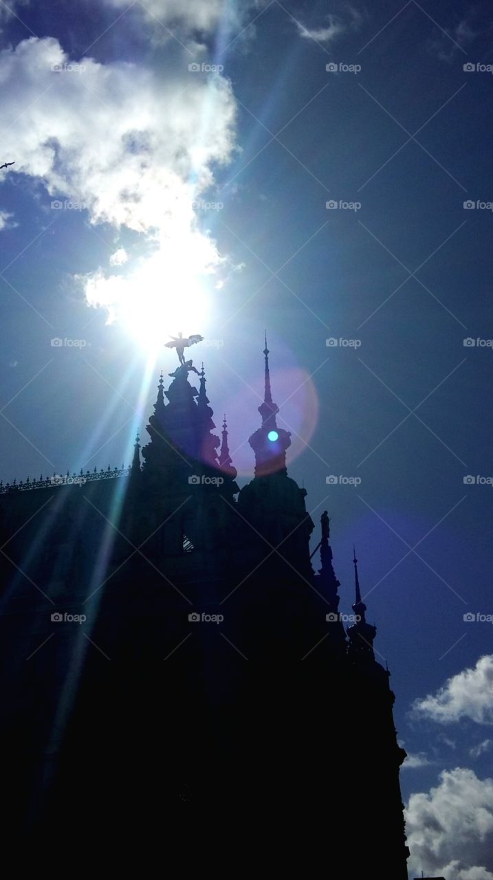 Angel statue, atop ornate spire, silhouetted against bright sun in blue sky.