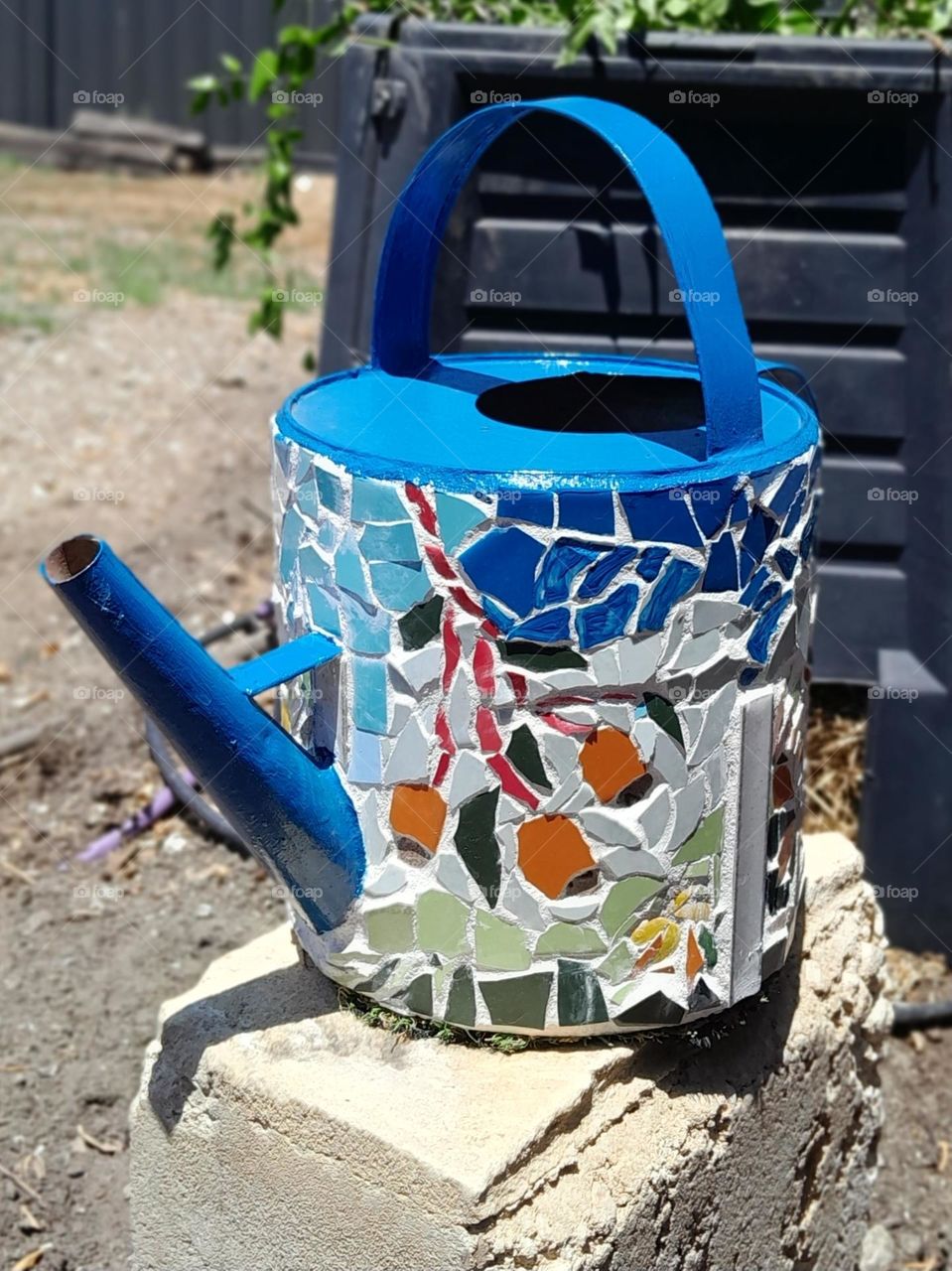 an old watering can, decorating and remade with mosaics and art, now beautifully standing in the middle of a garden.