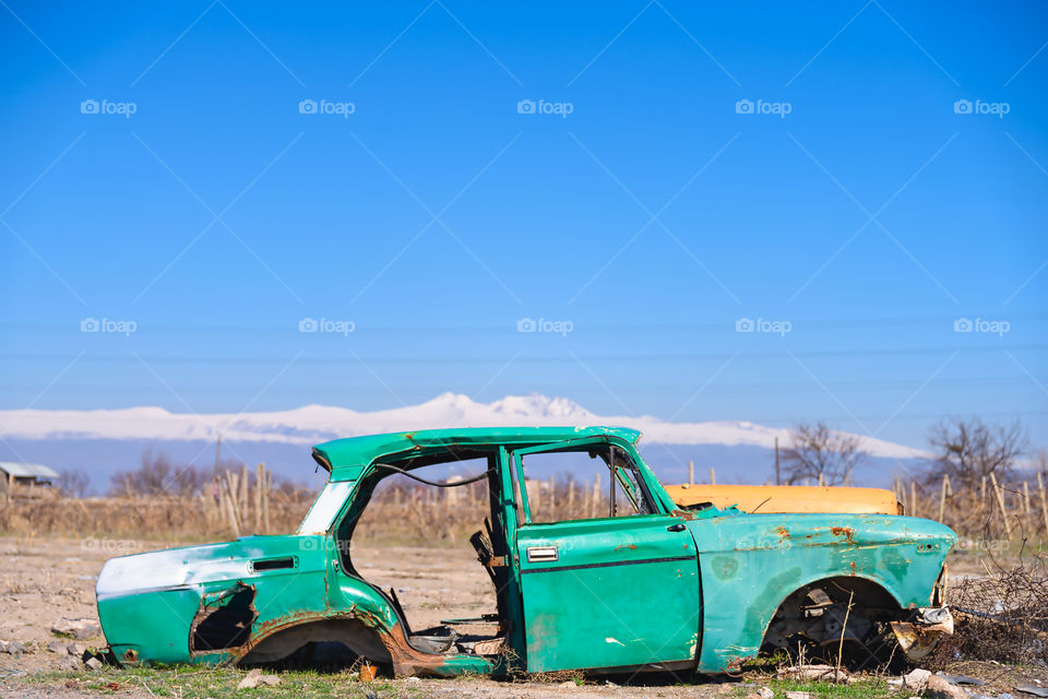 Abandoned and rusty wreck of an old green vintage Soviet Russian car in the middle of dry agricultural land with scenic snow-topped mountains and clear blue sky on the background in rural Southern Armenia in Ararat province on 4 April 2017.
