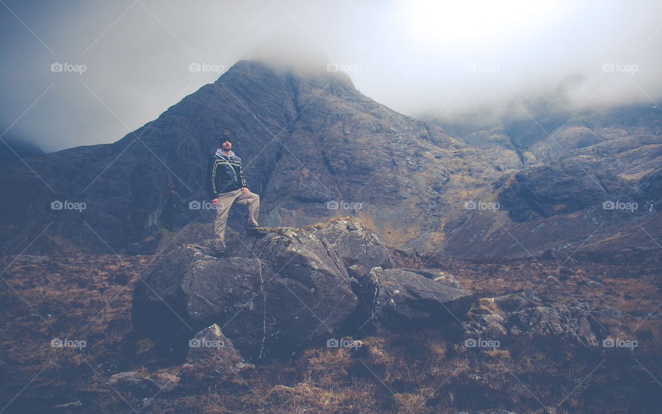 A man in warm clothes and woollen hat stands atop of some large boulders, surrounded by rugged, misty mountains 