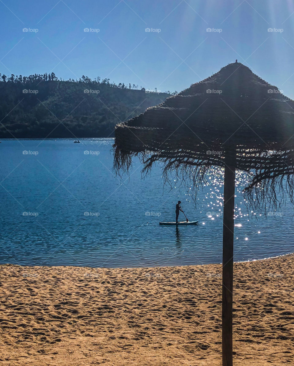 A paddle boarder glides along the aquamarine, river waters along the line of a sandy beach, with a parasol silhouetted against a clear blue sky