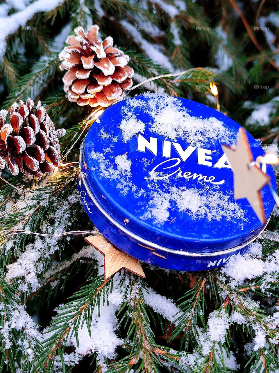 A blue jar of Nivea cream lies on the snow-covered branches of a spruce. Around the jar is a garland of pine cones, wooden stars and light bulbs. White snow on the lid of a jar of cream