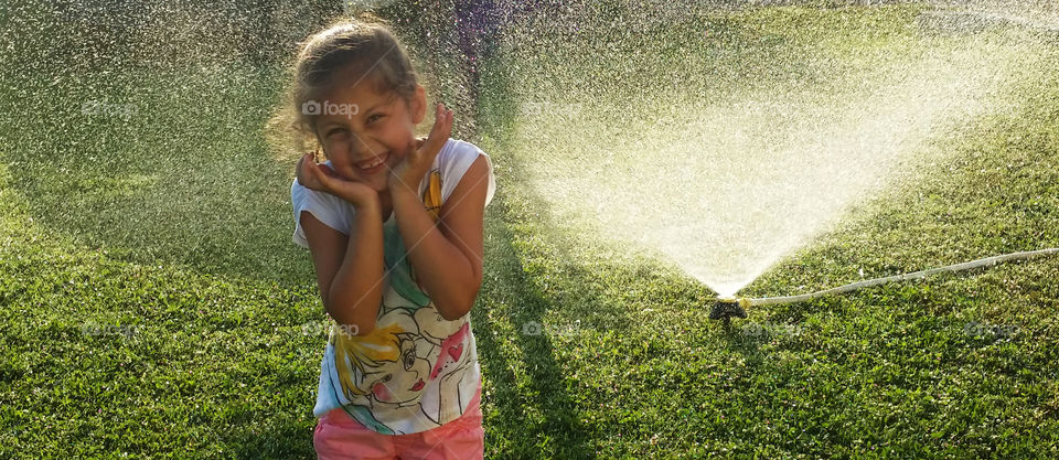 Happy girl standing near water sprinkler in garden