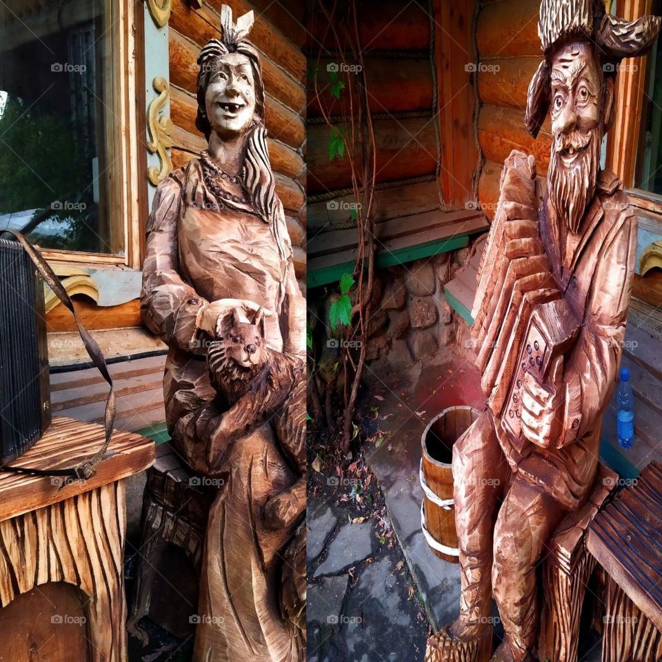 Happiness.  Two photos in one.  The first photo shows a wooden sculpture of a smiling grandmother sitting on a bench petting a cat.  In the second photo there is a wooden sculpture of a smiling grandfather who plays the button accordeon