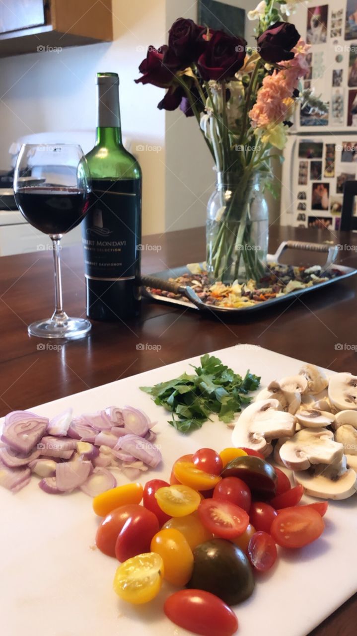 Preparing for a romantic evening with basil, onion, tomatoes, and mushrooms waiting to be cooked, red wine waiting to be drank, and fresh flowers. 