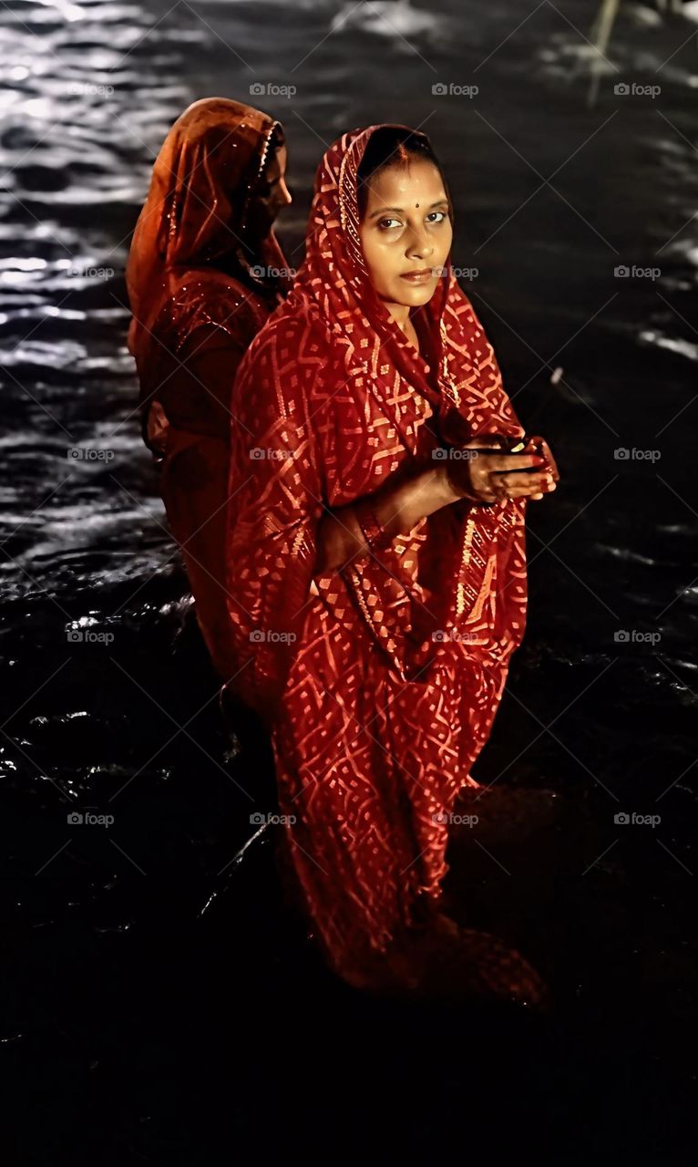 A powerful portrayal of Chhath Puja, this photograph captures two women in traditional red attire, immersed in water as they perform prayers with serene devotion under the night sky, embodying faith and cultural heritage.