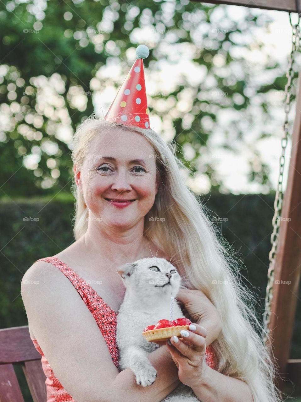 Portrait of a beautiful young blonde woman with a cone hat on her head, a tartlet with a candle and a gray thoroughbred cat in her arms,sits on a swing in the garden,vtd side close-up. Birthday celebration concept.