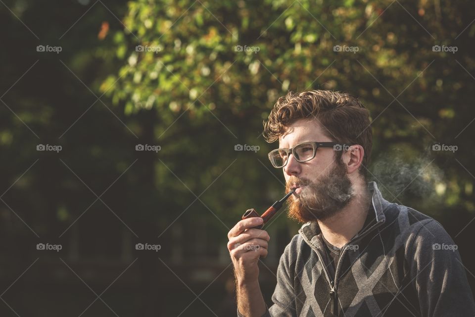 Hipster smoking a pipe outside. Young man smoking a pipe with trees behind 
