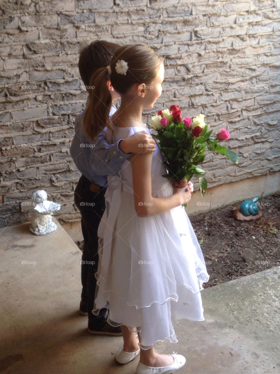 Two young Children stand close, viewed from rear side. The girl is wearing white, holding a bouquet of colourful roses, hair in a neat pony tail. They stand in an entrance way for photos. 