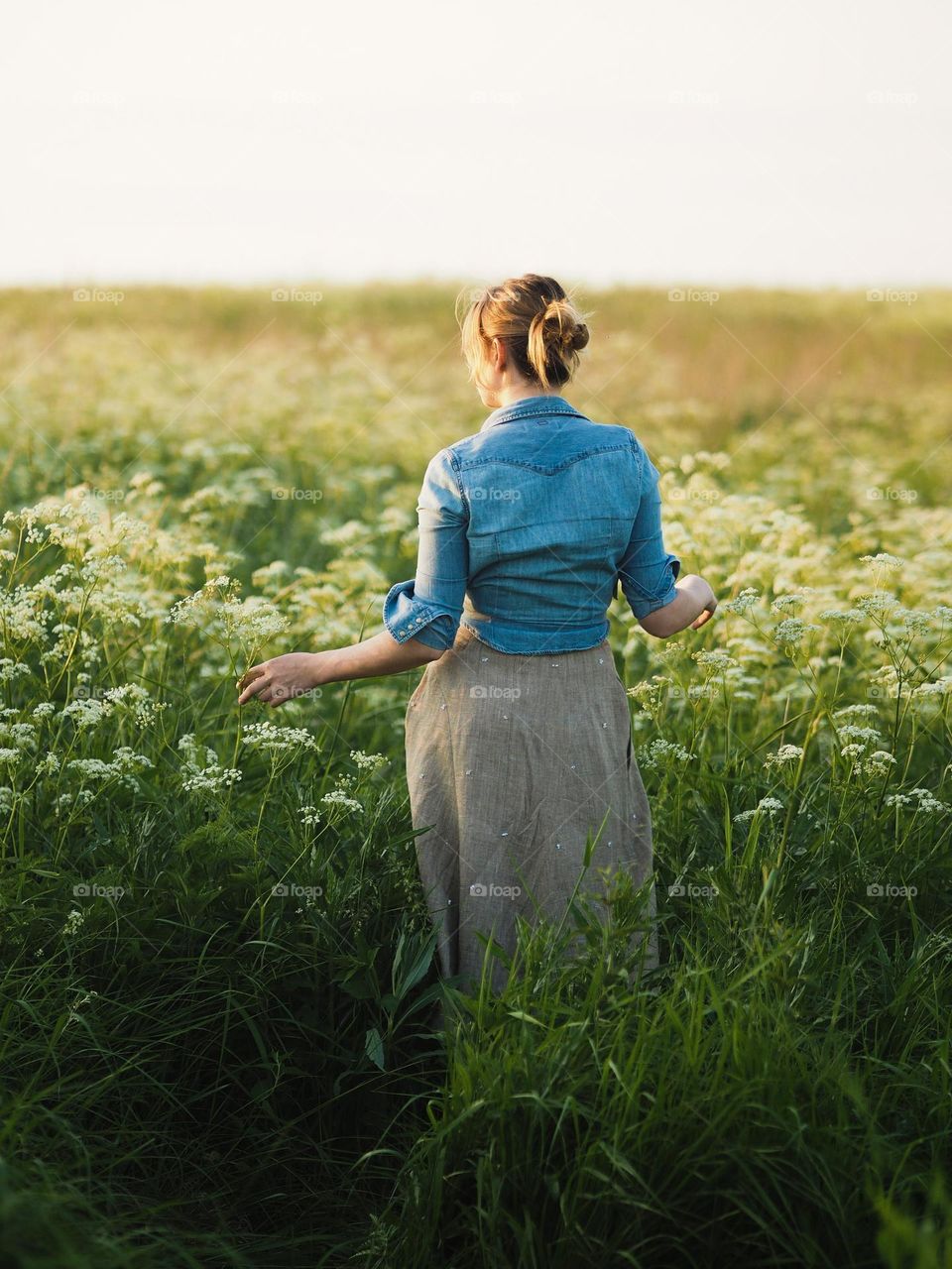 Young blonde woman walking in blooming field on a summer day in countryside, back view 