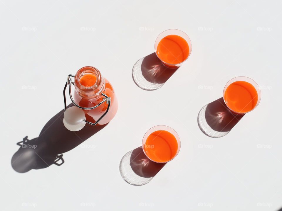 One small glass bottle and three mini glasses with orange juice stand on a white table with hard shadows from the sun, flat lay close-up. Concept summer drinks, light and shadows.