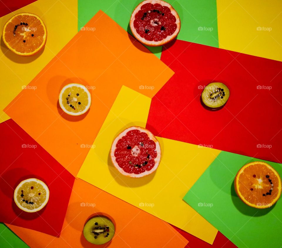This image features sliced fruits—grapefruit, oranges, lemons, and kiwis—arranged on a colorful background of red, yellow, green, and orange paper. The fruits have black seeds forming smiley faces, creating a playful and vibrant composition.