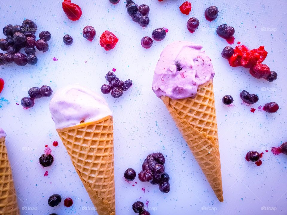 Two wildblueberry coconut ice-cream on waffle cones surrounded by wild blueberries,raspberries and blue sugar sprinkles.