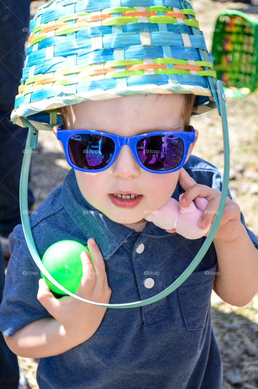 Close-up of a toddler boy with an Easter basket on his head and holding Easter eggs