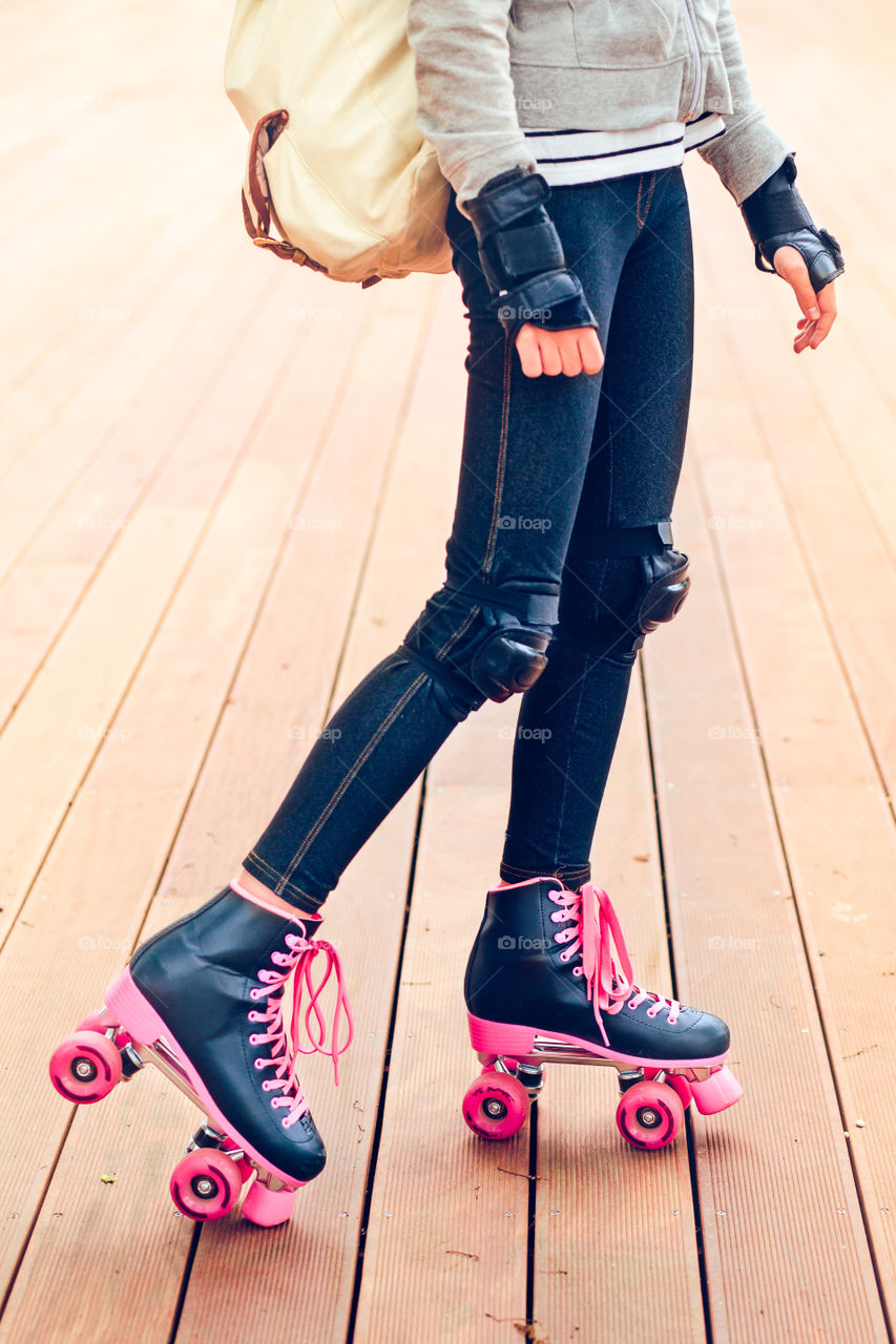 Close up of legs of teenage girl wearing jeans and pink black skates standing on wooden stage with backpack and protective sportswear