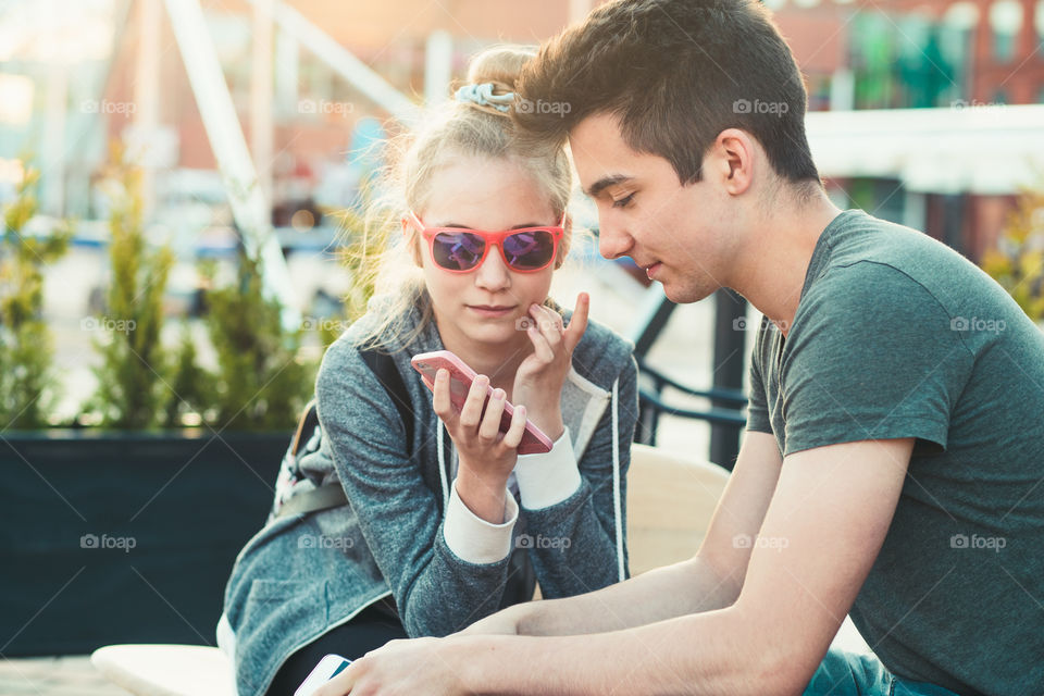 Couple of friends, teenage girl and boy, having fun using smartphones sitting in center of town, spending time together