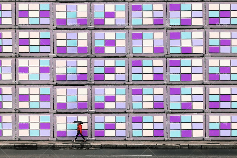 Tiny human walking with umbrella in front of a purple geometric wall background 