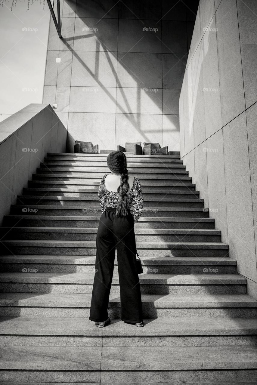 A fashionable girl stands on the steps of a building on the walls of which there is a beautiful play of light and shadow.