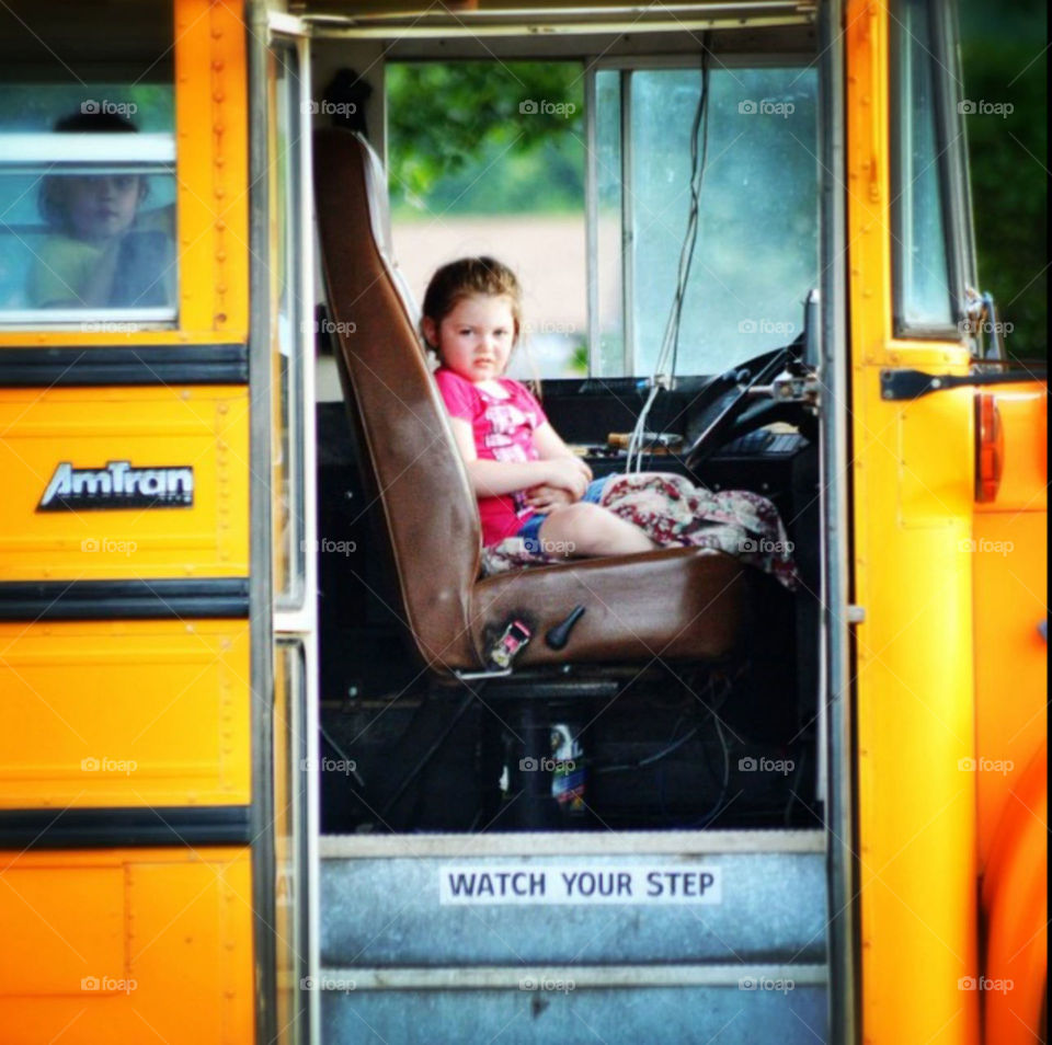 Intrigued child sitting in the bus driver's seat while another peeks out the window.