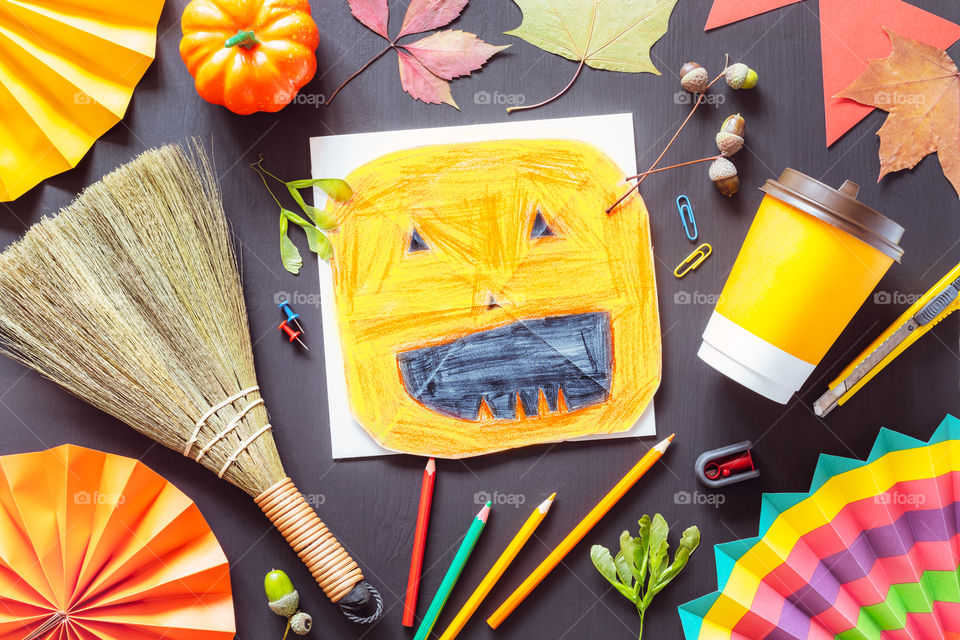 Autumn still life with children drawing of a halloween pumpkin, color pencils and autumn decorations on a black chalkboard. Art workplace for halloween creativity. Pre school lesson. Flat lay