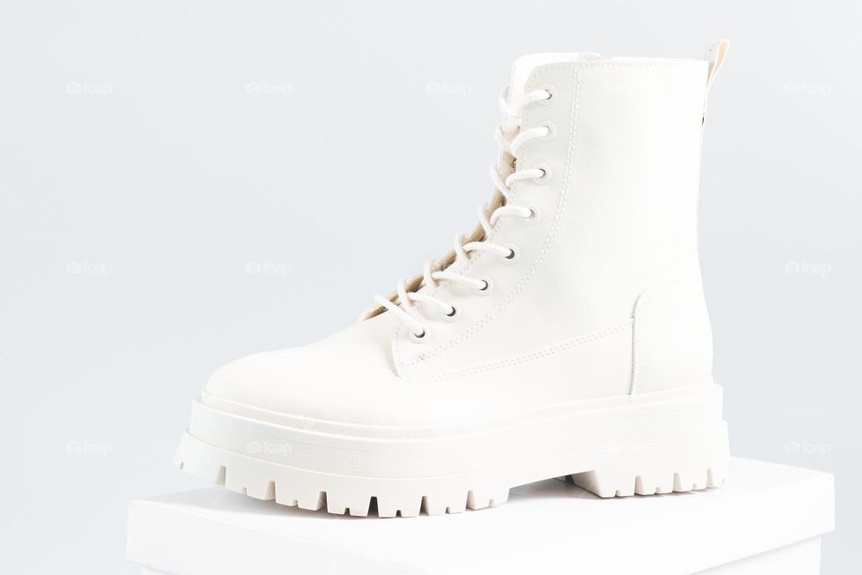 One white fashionable tractor-soled boot with lacing stands on a white stand on a white background, close-up side view. The concept of women's shoes, white on white.