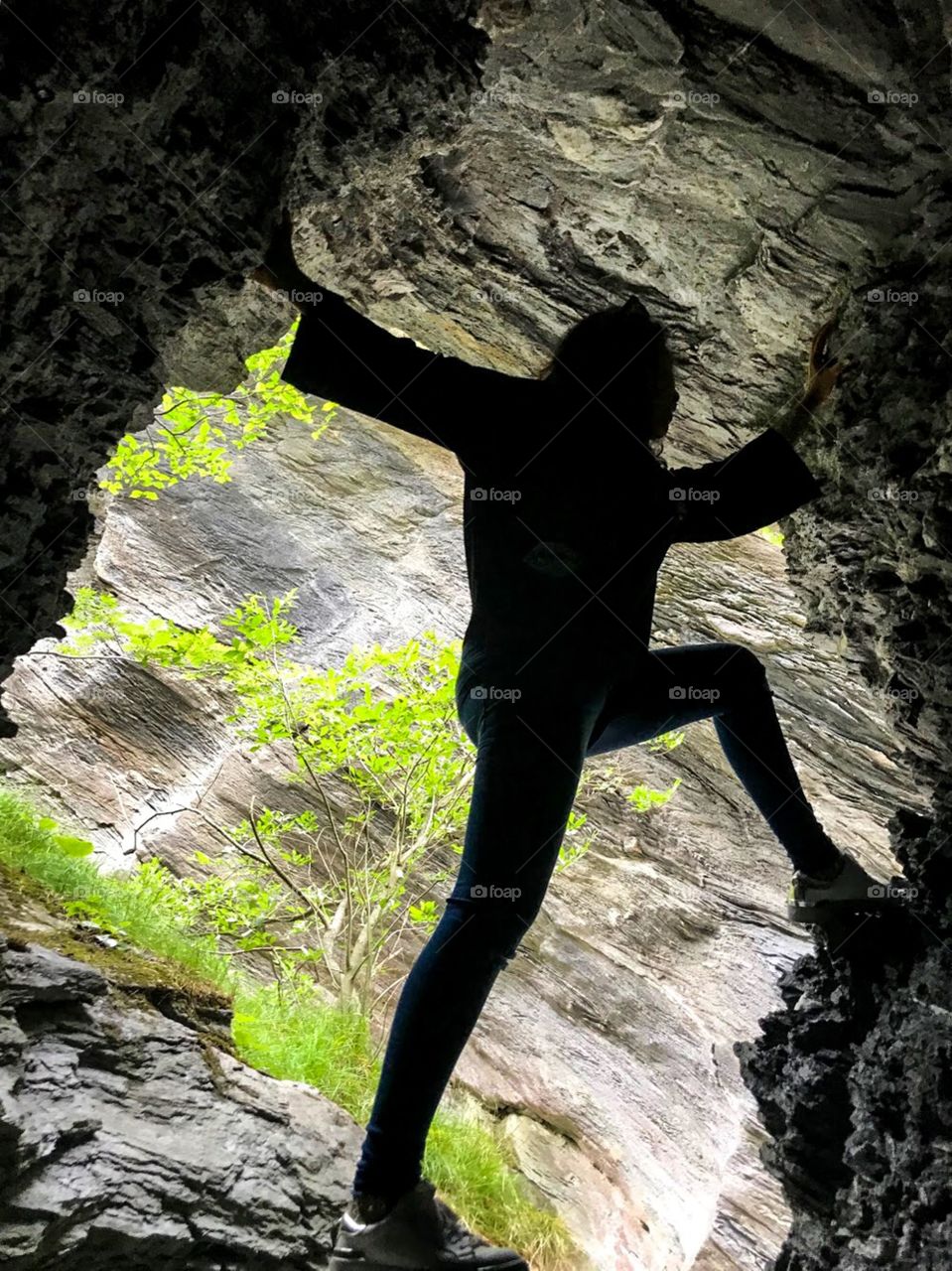 woman in black t-shirt, blue jeans, white shoes posing at void of rocky mountain . the cliffs are black and gray, and there are grass and some green trees