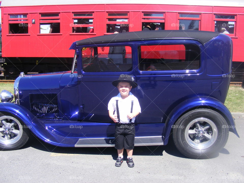 Young and Old. An excited young boy stands proudly in front of an old classic blue motor vehicle. Behind is a red carriage of an old steam train. It is Art Deco weekend in Napier NZ