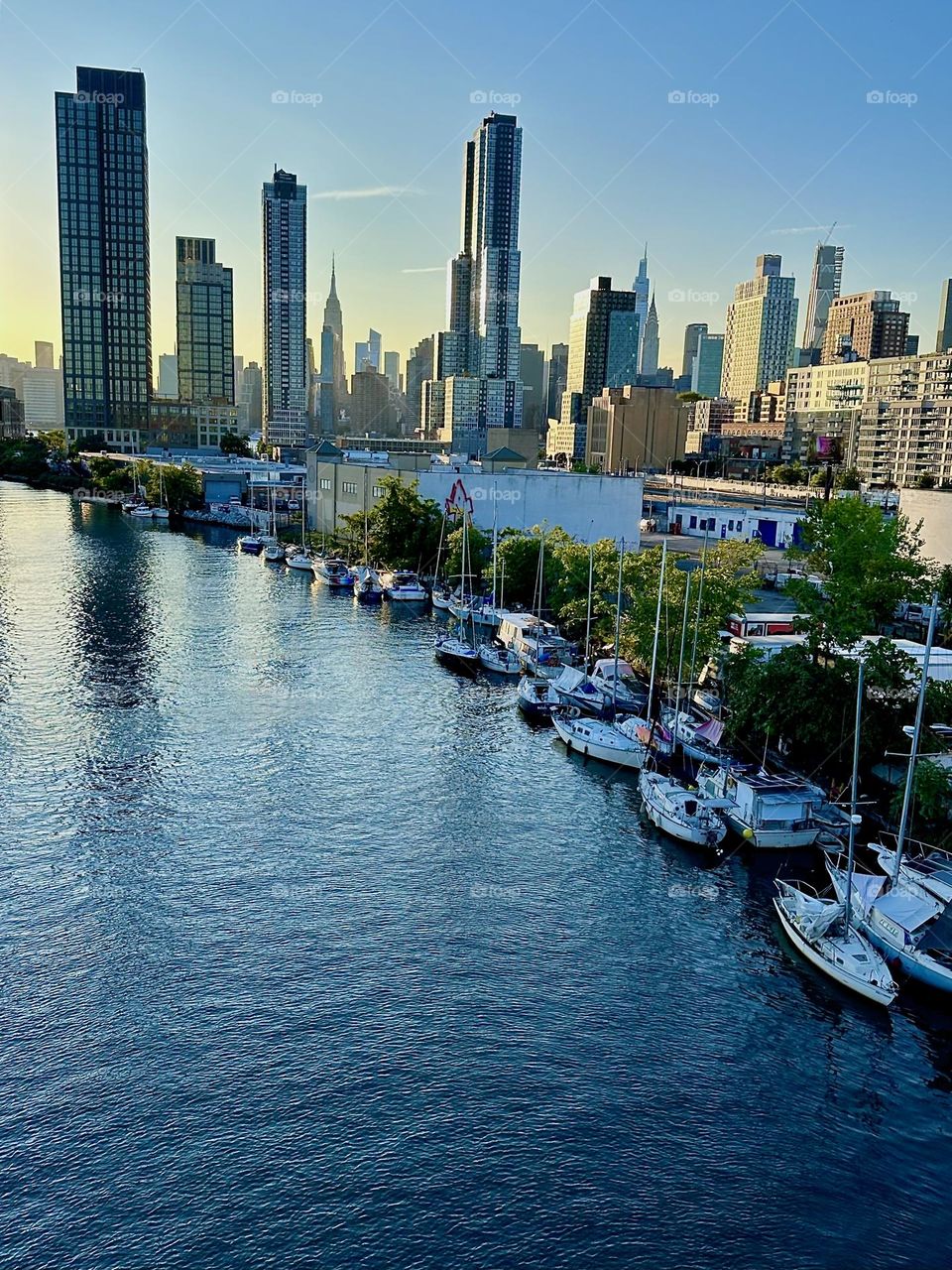 This is “Newtown Creek” with its variety of boats lined up side by side seen from the “Pulaski Bridge” that connects “Greenpoint”, Bklyn to LIC. In the distance “Manhattan” including the “Empire State Bldg” is visible. 2024. Hypnotic Productions