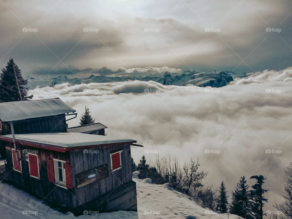 A wooden house on top of a mountain with red windows . A lot of clouds on the top of the mountain . Beautiful landscape 