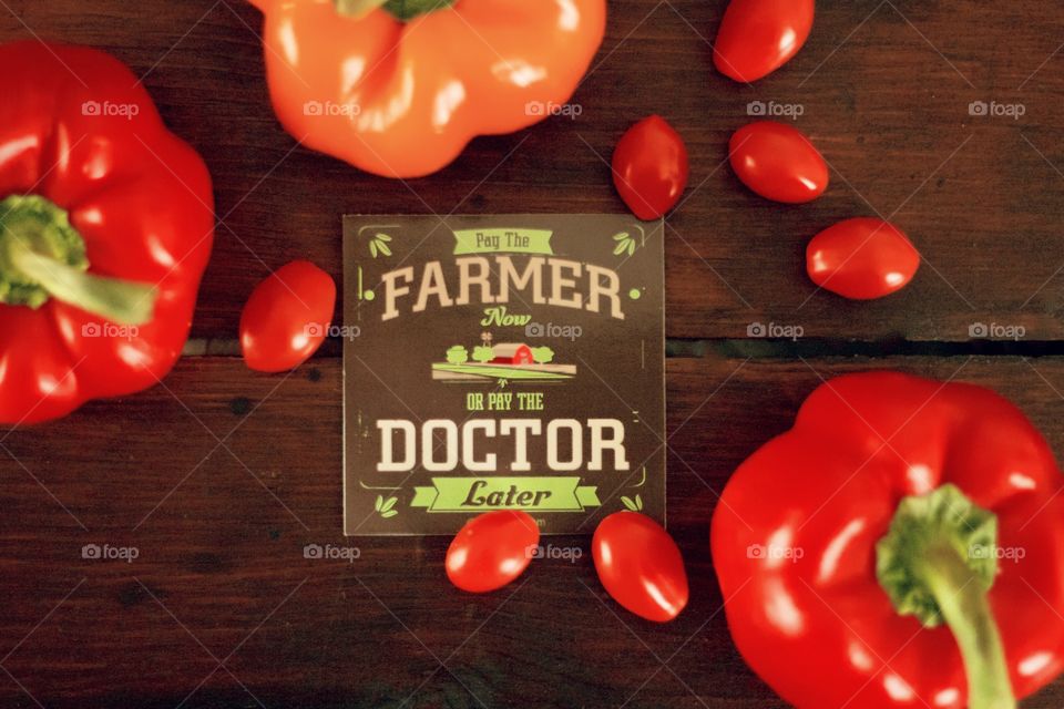 Flat lay of “Pay the Farmer Now or Pay the Doctor Later” sign with red and orange bell peppers and grape tomatoes on dark wooden surface