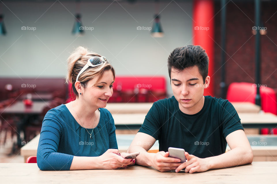 Woman and young man talking together and using mobile phones sitting by a table in cafe