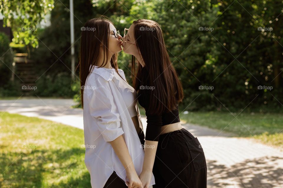 Two beautiful caucasian brunette sister girls stand in a public park and gently kiss each other on the lips, close-up side view. The concept of kissing people.