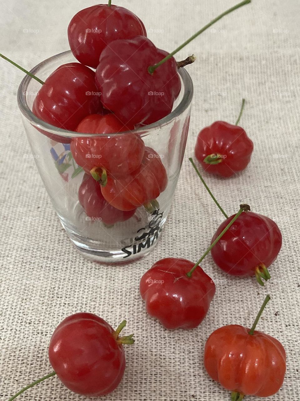 Pitangas naturais vermelhas dentro de recipiente e fora em forma decorativa. A composição está sobre uma toalha em cor crua.