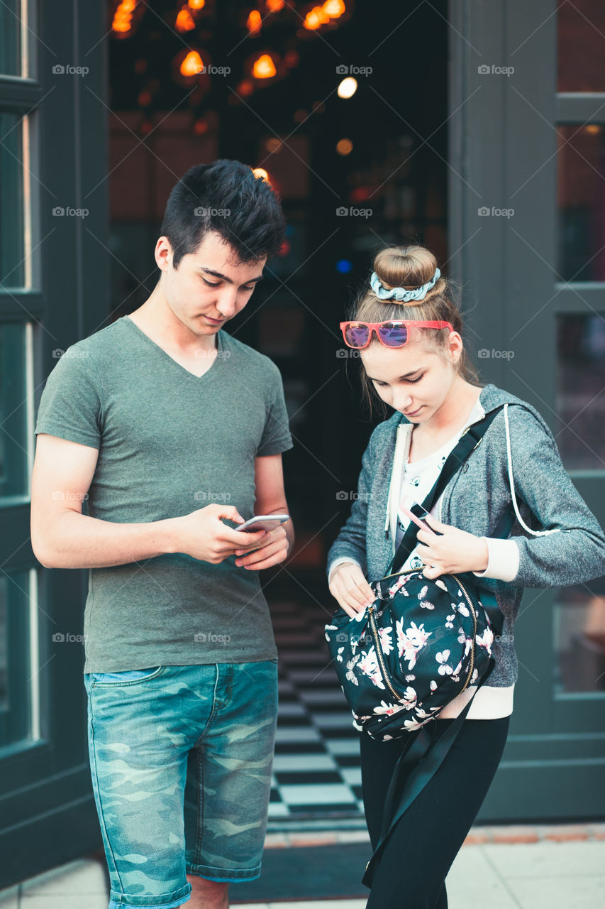 Couple of friends, teenage girl and boy,  using smartphones, talking together, standing on street in center of town, spending time together