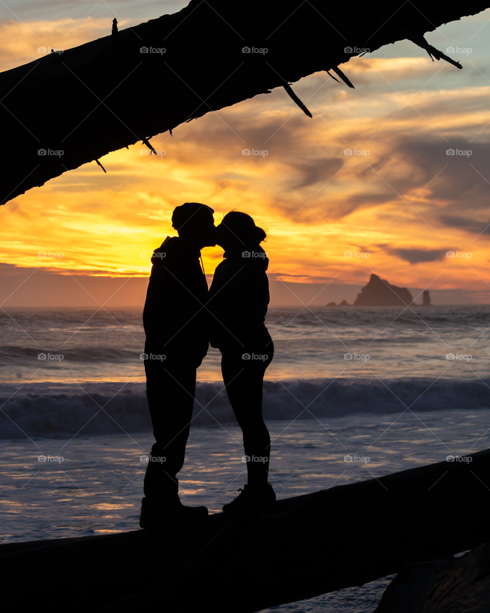 A silhouetted couple kissing, as a vibrant orange and yellow sunset bathes the beach and coast in golden light. 