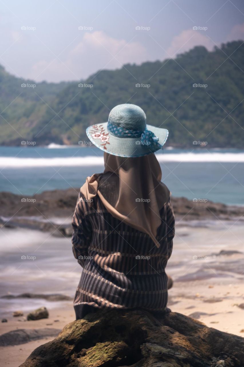 a woman sitting on a rock by the beach and wearing a polka dot hat