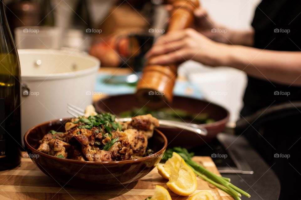 Grinding pepper over fresh vegetables in frying pan with bowl of fresh homemade chicken with lemon pieces on foreground and bottle of wine home cooking photography 